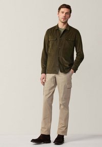 Camicia a maniche lunghe in velluto a coste verde scuro con due tasche sul petto, chiusura con bottoni, abbinata a pantaloni cargo beige e scarpe marrone scuro.