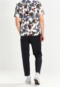 Camicia bianca a maniche corte con motivi floreali in rosso, blu e verde; abbinata a pantaloni scuri a sigaretta e sneaker grigie.