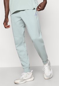 Gaiši zilas sporta bikses no mīksta auduma, ar sašaurinātu kāju dizainu un purpursarkanā izšuvuma logotipu uz sāniem.