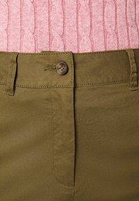 Un pantalon vert olive présente une texture lisse, une fermeture à bouton et des passants de ceinture. Au-dessus, un pull en tricot rose clair avec un motif en câble.