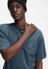 Sininen lyhythihainen T-paita, jossa pystysuuntaiset raidat, pyöreä pääntie ja teksturoitu merkki, jossa lukee "HUGO." Malli käyttää mustaa pipoa.