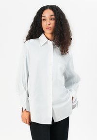 Witte oversized button-up shirt met een puntige kraag, lange mouwen en een enkele zak. De stof heeft een gladde textuur en een subtiele glans.