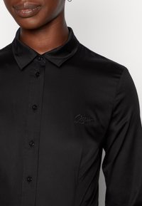 Schwarzes Langarmshirt mit spitzem Kragen, vorderen Knöpfen und gesticktem Logo. Glatte Textur mit figurbetontem Schnitt.