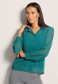 Türkisfarbene Bluse mit durchgehendem Knopfverschluss, spitzem Kragen und langen Puffärmeln. Getragen mit schwarz-weißen gemusterten Hosen.