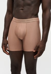 Άνδρας που φοράει ανοιχτό καφέ μποξεράκια Under Armour με υφασμάτινη υφή και ζώνη με την επωνυμία, στέκεται με τα χέρια χαλαρά δίπλα στο σώμα του.