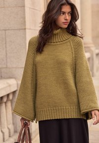 Khaki green oversized sweater with a high, ribbed turtleneck. Features wide, textured sleeves and a ribbed hem, paired with a dark skirt.
