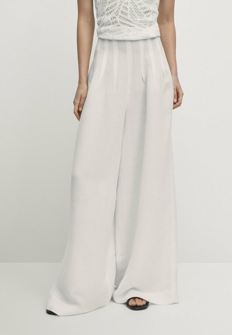 Personne portant un pantalon blanc taille haute à jambes larges et un haut en maille blanc sans manches, debout devant un fond clair uni.