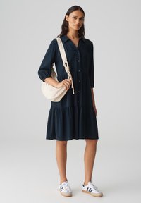 Abito blu navy con bottoni, maniche a tre quarti e gonna a strati; abbinato a una borsa a spalla crema e sneakers bianche con dettagli blu.