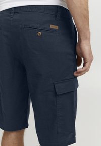 Shorts cargo bleus marine pour hommes en tissu texturé. Les caractéristiques incluent une poche latérale, une poche arrière avec bouton et une étiquette sur la ceinture.