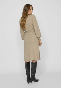 Vestito lavorato a maglia beige con cintura in vita, maniche lunghe a palloncino e silhouette al ginocchio, abbinato a stivali neri con tacco alti fino al ginocchio.