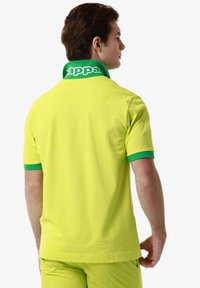 Polo giallo con colletto e polsini verdi, con logo del marchio sul colletto, che presenta una texture liscia e un design informale.