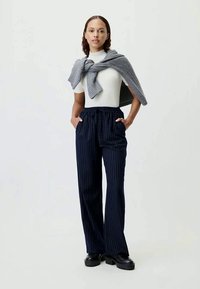 Mujer vestida con una blusa blanca de manga corta, suéter gris drapeado sobre los hombros, pantalones de pierna ancha de rayas navys y zapatos negros, de pie con las manos en los bolsillos.
