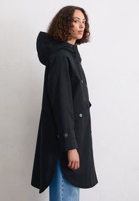 Schwarzer Kapuzenmantel mit geschwunghem Saum, aus glattem Stoff gefertigt. Verfügt über Knopfverschlüsse und Knöpfe an den Bündchen. Getragen mit hellblauen Jeans.