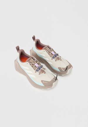 Zapatillas de trail ligeras, en tonos beige y gris, con un upper texturizado, acentos en púrpura y aqua, y suela de goma con tacos de tracción prominentes.