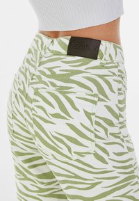 Vita denimbyxor med ett grönt zebra-randigt mönster, som har en brun läderlogotyp på midjan och två bakfickor.