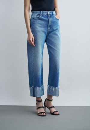 Mujer con vaqueros cropped azul claro con puños y bajos deshilachados combinados con sandalias negras de tacón alto con tiras sobre fondo liso.