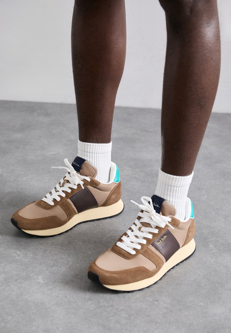 Baskets marron et beige avec des lacets blancs, en cuir et en daim, un accent bleu-vert au niveau du talon, et une semelle en caoutchouc crème.