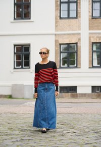 Femme portant un pull rayé noir et rouge, une longue jupe en denim, des ballerines noires et des lunettes de soleil marchant sur une rue en pavés près d'un bâtiment blanc.