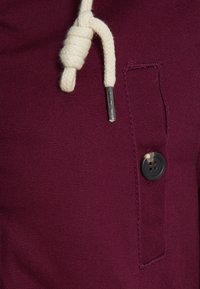 Mörkröd tyg med en strukturerad yta, prydd med en svart knapp och en krämfärgad dragsko. En anmärkningsvärd fickdetalj syns.