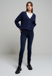 Jūras zilas adītas kardigāns ar V veida kakla izgriezumu, lielām pogām un ribotām piedurknēm, apvienots ar tumšām šaurām džinsām un melniem šņorējamajiem apaviem.