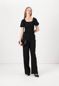 Crni smockani crop top s rastegnutim rukavima, uparen s visokim crnim hlačama; drži malu crnu torbicu. Glatka tekstura, minimalni naglasci.