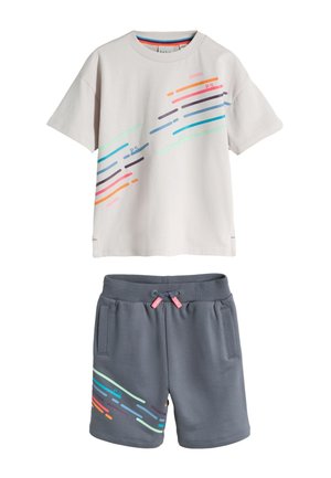 Jongen staat, draagt een lichtgrijs T-shirt met kleurrijke strepen, donkergrijze korte broek met vergelijkbare strepen, witte sokken en witte sneakers.