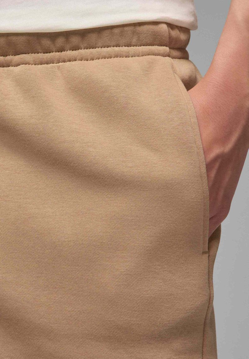 Pantalons de survêtement beige au toucher lisse, avec une poche latérale et une taille élastique. L'angle en gros plan met en valeur le tissu et les coutures.