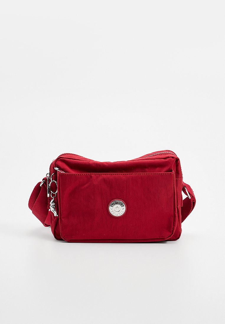 Röd tyg crossbody-väska med framficka med dragkedja, justerbar axelrem och runt silverlogotyp-emblem på en enfärgad bakgrund.