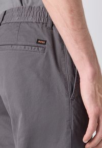 Närbild på en person som bär grå shorts med elastisk midjeresår och en liten "BOSS"-etikett ovanf ör bakfickan på höger sida.