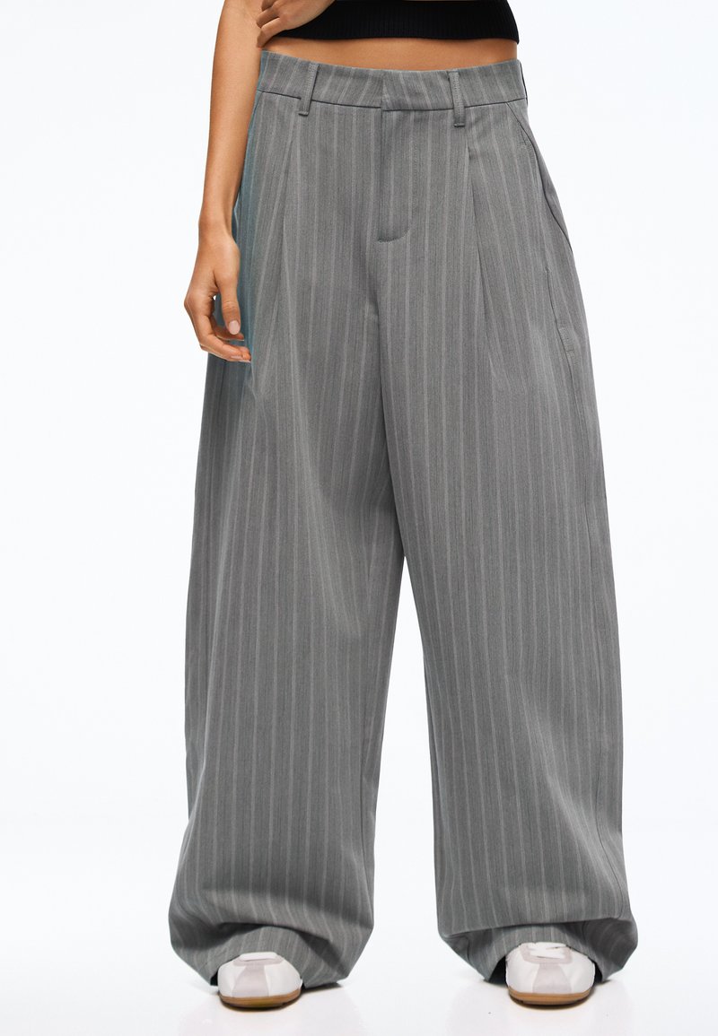 Personne portant un pantalon large gris à fines rayures et des baskets blanches, debout avec une main détendue le long du corps.