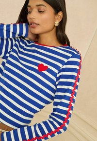 Blau-weiß gestreiftes Langarmshirt mit einer roten Herz-Applikation auf der Brust und rotem, wellenförmigem Besatz entlang der Ärmel. Gemütliches Baumwollmaterial.