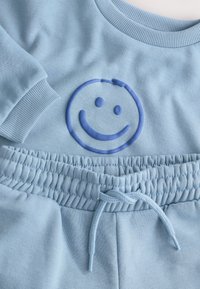 Világoskék pulóver bordázott mandzsettákkal, kiemelkedő kék mosolygós arcmintával. Hozzáillő, állítható derékpántos melegítővel.