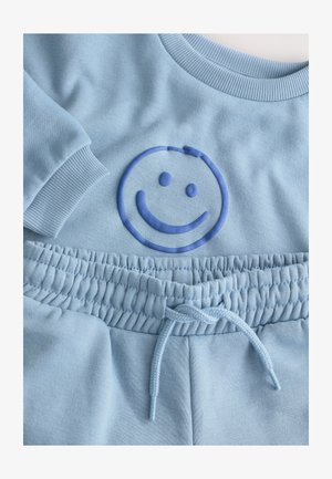 Svetlo modra jopa z rebrastimi manšetami, z dvignjenim modrim nasmeškom. V kompletu s primernimi hlačami za tek z nastavljivim pasom.