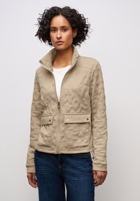 Beige vadderad jacka med hög krage, dragkedjestängning och två framfickor. Strukturerad tyg med ett subtilt mönster, matchad med blå jeans.
