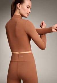 Bruine lang-sleeve cropped top gecombineerd met high-waisted leggings, beide gemaakt van gladde stof. Bevat minimale branding op de tailleband.