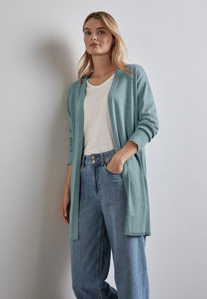 Cardigan verde chiaro con apertura frontale, polsini a coste e vestibilità comoda, abbinato a una maglietta bianca e jeans blu a vita alta.
