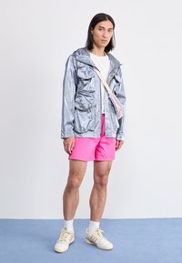 Sølvreflekterende jakke med flere frontlommer, parret med lyserøde shorts, hvide sokker og beige sneakers med gyldne detaljer.