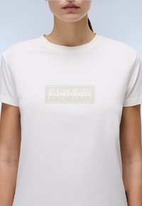 Hvid bomulds T-shirt med korte ærmer, med et rektangulært beige logo "NAPAPIJRI geographic" på brystet. Glat tekstur.