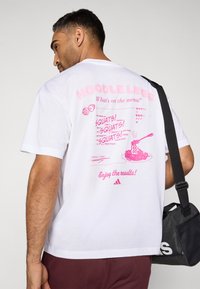 Biele bavlnené tričko s ružovým grafickým nápisom na chrbte. Dizajny zahŕňajú "NOODLE LEGS", pripomienky na cvičenie a ilustráciu cestovín.