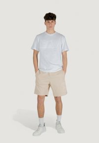 T-shirt bianca a maniche corte con logo "EA7" in rilievo abbinata a pantaloni corti beige con stampa del logo. Il modello indossa sneakers bianche e calze.