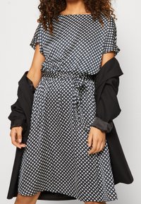 Robe bleue avec motif géométrique blanc, manches courtes, taille élastique et ceinture à nouer. Associée à une veste noire. Texture de tissu lisse.