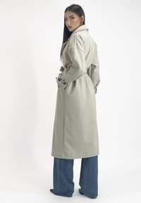 Cappotto lungo grigio chiaro con cintura, polsini bottonati e colletto strutturato. Indossato sopra jeans a gamba larga blue. Texture liscia.