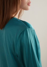 Teal satinblus med en mjuk textur, breda axlar och en avslappnad passform. Tyget faller mjukt och framhäver en flytande design.