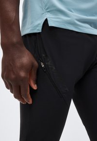 Κοντινό πλάνο ενός ατόμου που φοράει μαύρο αθλητικό παντελόνι με τσέπη με φερμουάρ και γνωρίσματα ανοιχτό μπλε τελειώματος πουκάμισου πάνω από τη μέση.