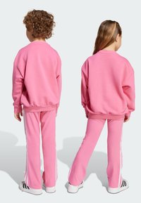 Ružový látkový sweatshirt a zodpovedajúce nohavice s bielymi bočnými pruhmi. Voľný strih, okrúhly výstrih a rebrované manžety. Hladká textura, ležérny štýl.
