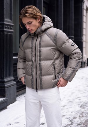 Jeune homme en doudoune grise et pantalon blanc debout dans une rue urbaine enneigée, les mains près des poches.