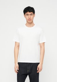 Νεαρός άνδρας με κοντά μαύρα μαλλιά, φορώντας ένα απλό λευκό κοντομάνικο μπλουζάκι και σκούρα τζιν, στέκεται μπροστά σε λευκό φόντο.