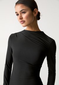 Abito lungo nero a maniche lunghe con scollatura drappeggiata e texture liscia. Il tessuto appare aderente, mettendo in risalto dettagli sottili sulle spalle.
