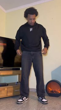 Felpa nera con un logo bianco, jeans scuri e sneaker Nike in bianco e nero. In piedi in una stanza con una TV e un camino.