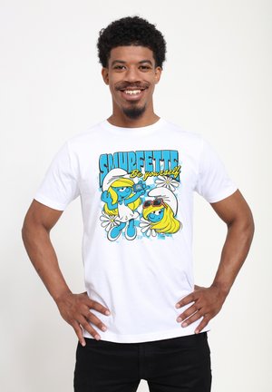 Lächelnder Mann mit kurzen lockigen Haaren, der ein weißes T-Shirt mit zwei Schlumpfine-Figuren und dem Text „Sei du selbst“ auf einem einfarbigen Hintergrund trägt.